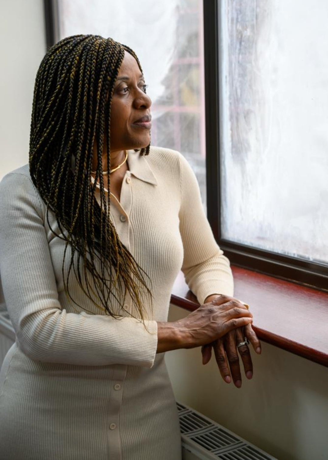 A woman with long dark braids wearing a cream-colored long-sleeve top and skirt, leaning against a window ledge in an indoor setting.